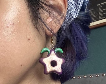 May include: A close-up of a single earring. The earring is a star shape with a hole in the center. The star is purple with gold accents and green curved accents. The earring is attached to a gold hook.