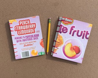 May include: Two small notebooks with covers made from repurposed juice boxes. The left notebook has a pink cover with the text "Peach Strawberry Lemonade" and a barcode. The right notebook has a purple cover with the text "Del Monte Quality te fruit" and a picture of a peach and strawberry.