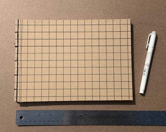 May include: A brown paper journal with a grid pattern and black lines. The journal is closed and has a black spine. A white marker is lying next to the journal. A ruler is in the bottom left corner of the image.