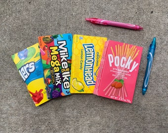 May include: A close-up of five different candy and snack packages. The packages are arranged horizontally from left to right. The packages are:  'Lakers' candy, 'Mike and Ike Mega Mix' candy, 'Lemonhead' candy, and 'Pocky Strawberry Cream Covered Biscuit Sticks'.