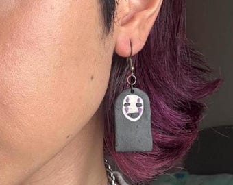May include: A pair of gray clay earrings with a white face and purple eyes. The earrings are shaped like a rectangle and have a silver hook.