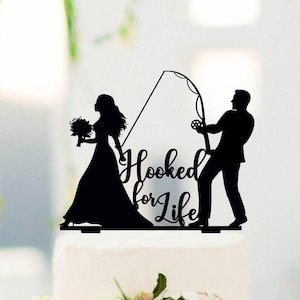 Puede incluir: Silueta negra de una novia y un novio con una caña de pescar y el texto "Hooked for Life" en un pastel de bodas blanco.
