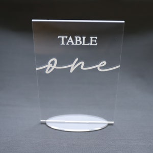 May include: Clear acrylic table number with a white base. The number "one" is written in a cursive font.