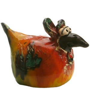 May include: A ceramic chicken figurine featuring a glossy finish with a vibrant colour palette of orange, yellow, red, and green. The bird has a dark brown head, prominent white eyes with black pupils, a pointed beak, and a raised tail section.