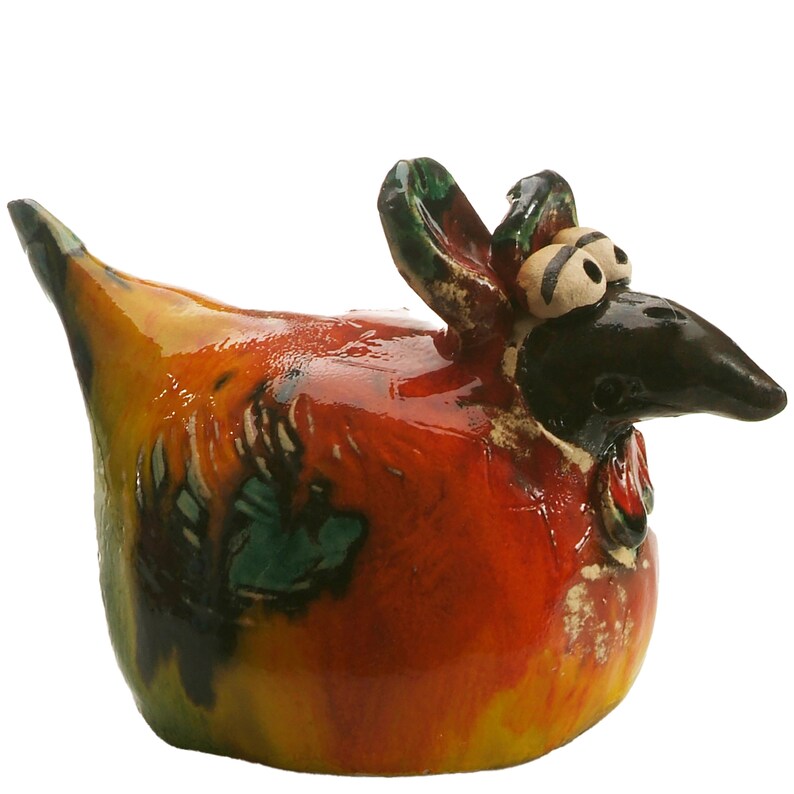 May include: A ceramic chicken figurine featuring a glossy finish with a vibrant colour palette of orange, yellow, red, and green. The bird has a dark brown head, prominent white eyes with black pupils, a pointed beak, and a raised tail section.