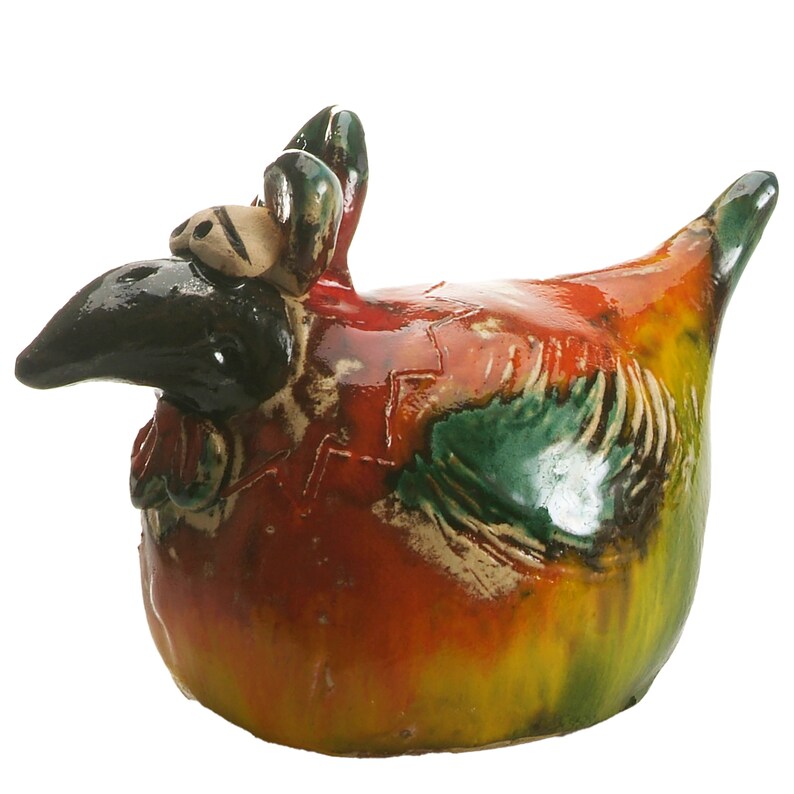 May include: A ceramic chicken figurine featuring a glossy, multi-coloured glaze in shades of red, yellow, and green. The sculpture has a prominent black beak, large eyes, and a textured wing detail, with a rounded body and a pointed tail.