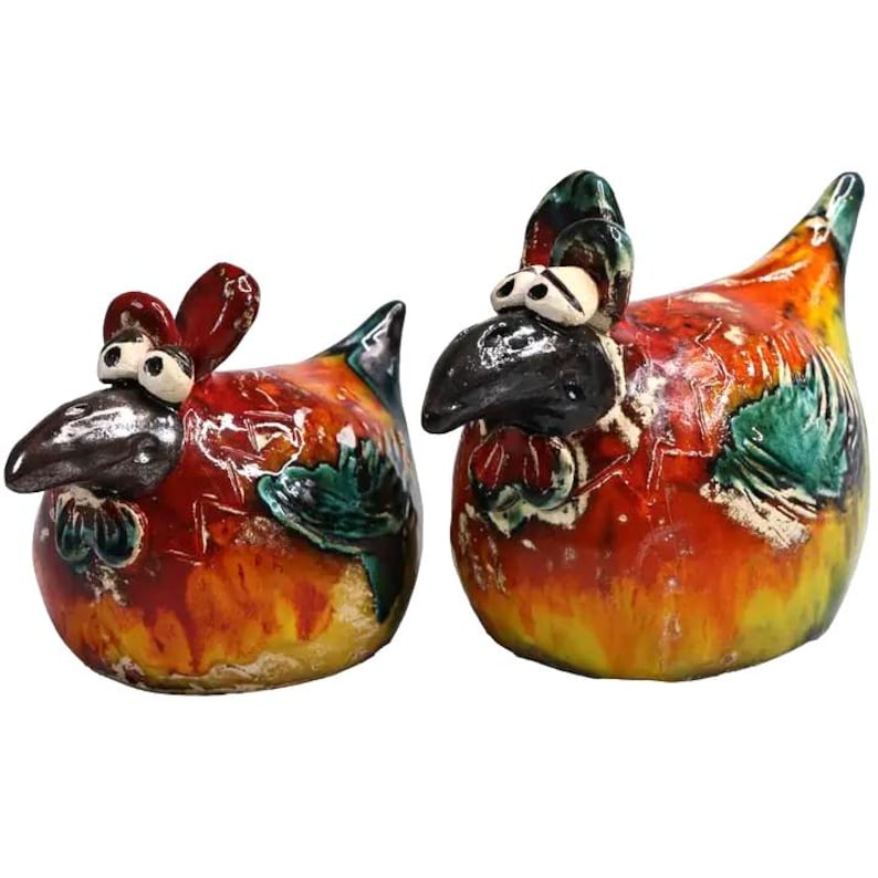 May include: Two ceramic chicken figurines featuring a glossy, multi-coloured glaze in shades of red, orange, yellow, and green. Each bird has a dark, textured beak, prominent white eyes with black pupils, and a rounded, plump body shape.
