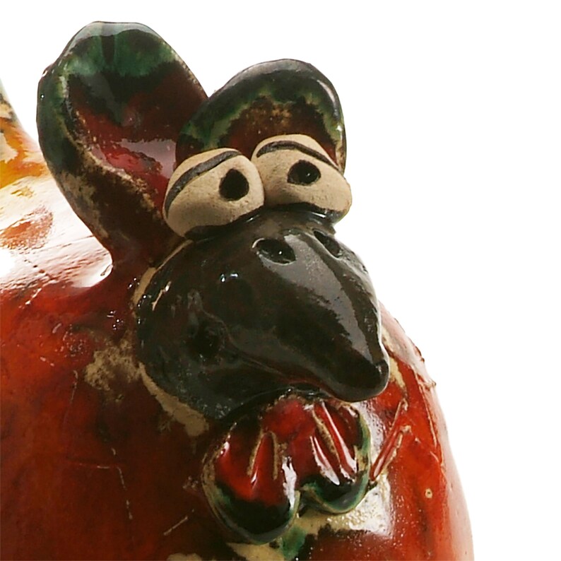 May include: A ceramic chicken figurine featuring a glossy red body with dark brown and green accents. The head has prominent white eyes with black pupils, a dark brown beak, and a red wattle. The surface has a smooth, reflective glaze finish.