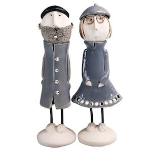 May include: A pair of ceramic figurines of a man and woman. The man is wearing a blue coat and a black hat. The woman is wearing a blue dress and a gray hat. Both figurines have white faces and black eyes.
