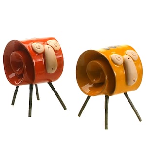 May include: Two ceramic animal figurines with glossy orange and yellow spiral-shaped bodies. Each figure features a light beige face with curled horns and stands on three thin, dark, textured metal legs. The designs are whimsical and abstract.