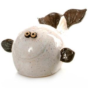 May include: A white ceramic fish figurine with brown fins and a speckled glaze. The fish has large, round eyes and a smiling mouth.
