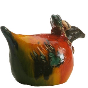 May include: A ceramic bird figurine with a glossy finish, featuring a rounded body painted in vibrant shades of orange, yellow, and deep green. The sculpture has a dark brown head, a pointed tail, and textured wing details on its sides.