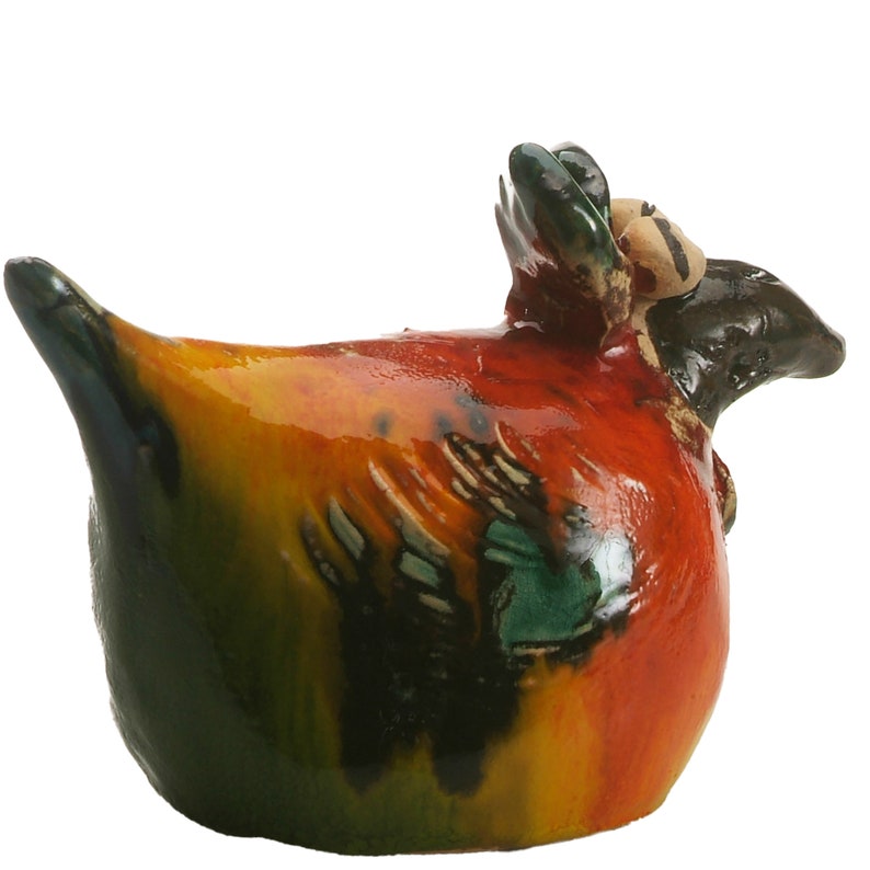 May include: A ceramic bird figurine with a glossy finish, featuring a rounded body painted in vibrant shades of orange, yellow, and deep green. The sculpture has a dark brown head, a pointed tail, and textured wing details on its sides.