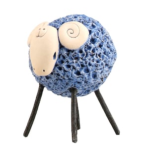 May include: A ceramic sheep figurine featuring a round, textured blue body with a smooth, off-white face and curled horns. The sculpture stands on four thin, dark brown, stick-like legs that provide a stark contrast to the porous blue wool texture.