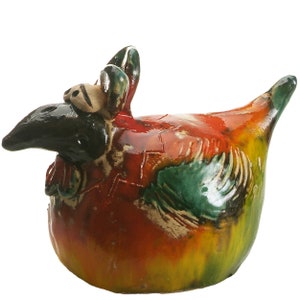 May include: A ceramic chicken figurine featuring a glossy, multi-coloured glaze in shades of red, yellow, and green. The sculpture has a black head, a prominent beak, and textured wing details, with a rounded body and a pointed tail extending upwards.