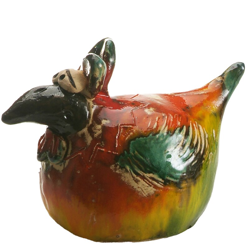 May include: A ceramic chicken figurine featuring a glossy, multi-coloured glaze in shades of red, yellow, and green. The sculpture has a black head, a prominent beak, and textured wing details, with a rounded body and a pointed tail extending upwards.