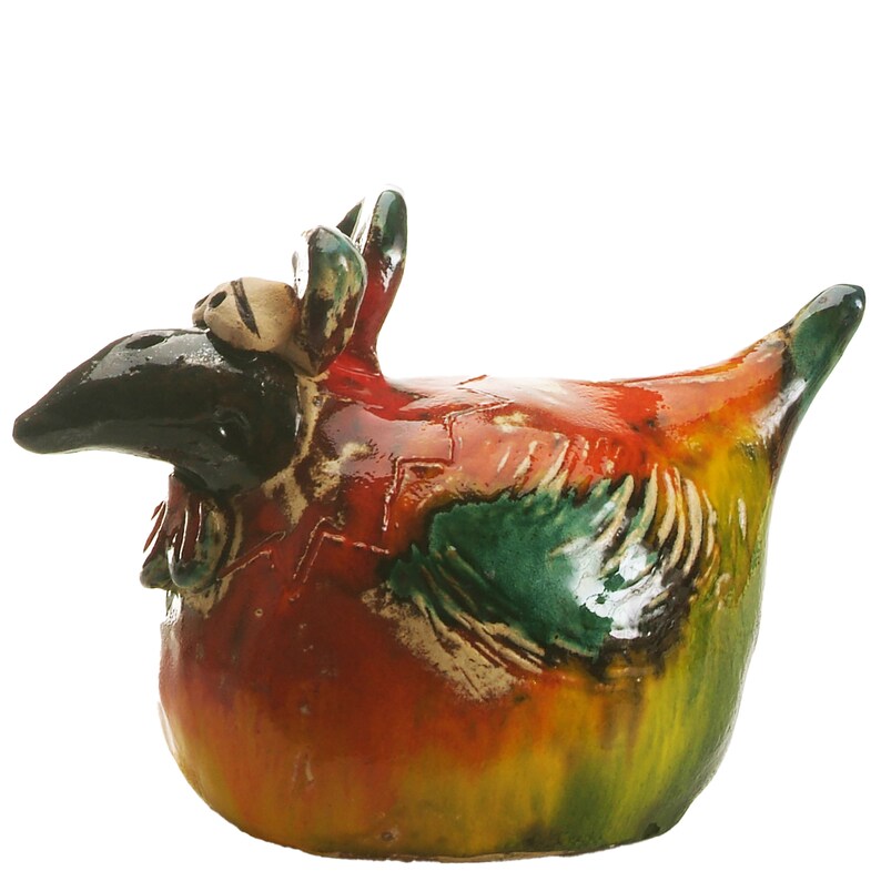 May include: A ceramic chicken figurine featuring a glossy, multi-coloured glaze in shades of red, yellow, and green. The bird has a prominent black beak, large white eyes with black pupils, and a textured wing detail on its side, all finished with a smooth, reflective surface.