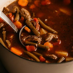 May include: A close-up of a pot of beef stew with a silver ladle. The stew contains chunks of beef, green beans, carrots, potatoes, and tomatoes in a rich, brown broth. The pot is off-white.