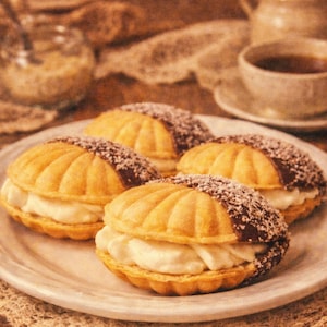 Op de afbeelding: Vier schelpvormige gebakjes met een crèmevulling en chocolade-kokos topping op een wit bord. Een kopje thee en een pot suiker staan op de achtergrond. De gebakjes hebben een goudbruine kleur.