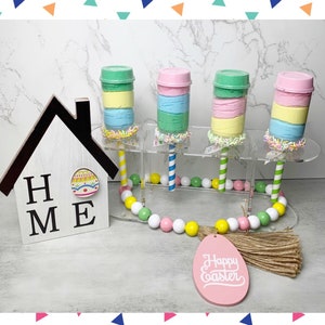 May include: Four clear plastic containers filled with layers of pastel-colored slime, topped with colorful sprinkles. The containers are on a clear acrylic stand with a beaded garland and a wooden sign that says "HOME" with a painted Easter egg. A pink wooden egg with the text "Happy Easter" is also included.