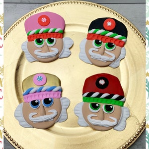 May include: Four nutcracker ornaments with different colored hats. The ornaments are on a gold plate. The hats are pink, black, gold, and red. Each ornament has a candy cane on the hat.