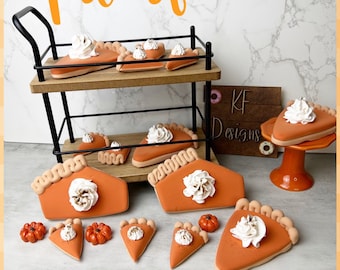 May include: A collection of orange pumpkin pie slice decorations with tan crust edges and white whipped cream dollops. The set includes various sizes displayed on a two-tier black metal stand, a small orange pedestal, and a flat surface with mini pumpkins.