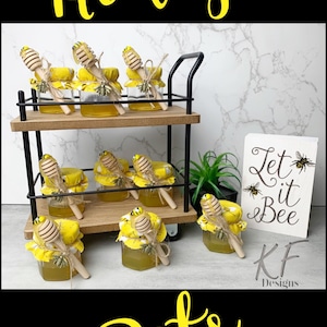 May include: A set of small glass jars filled with honey, decorated with yellow and black bee-themed fabric and wooden honey dippers. The jars are displayed on a black metal cart with wooden shelves. A sign reads "Let it Bee" with a bee design. Honey Pots.
