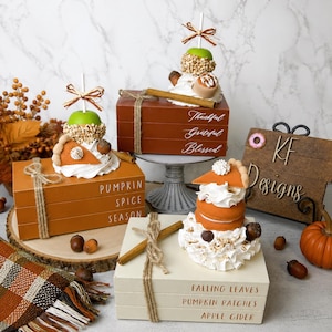 May include: Three stacks of decorative faux books in orange, brown, and cream colors, each tied with twine. The books feature text including Pumpkin Spice Season, Thankful Grateful Blessed, and Falling Leaves Pumpkin Patches Apple Cider.