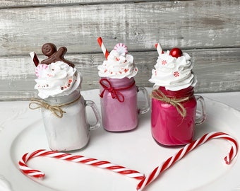 May include: Three glass mason jar mugs filled with white, light pink, and dark pink liquid, topped with white faux whipped cream, red and white striped straws, and small candy decorations. A red and white candy cane lies in front of the jars.