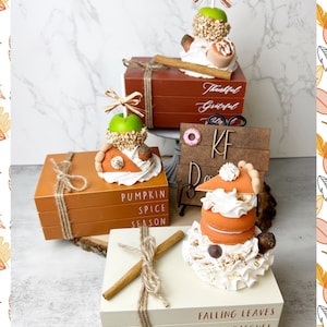 May include: Three stacks of books with fall-themed decorations. The top of each stack has a different fall-themed treat. The first stack has a caramel apple, the second stack has a pumpkin pie, and the third stack has a pumpkin spice latte. The books are decorated with twine and cinnamon sticks. The books have the following text: "Thankful Grateful", "Pumpkin Spice Season", and "Falling Leaves Pumpkin Patches Apple Cider".