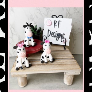 May include: Three small, hand-painted, ceramic cow figurines. The cows are white with black spots and have pink noses. The cows are sitting on a wooden shelf with a sign that reads "KF Designs".