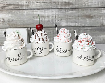 May include: Four white ceramic mugs arranged on a white platter, each topped with faux whipped cream and a black and white striped straw. The mugs feature black cursive text reading noel, joy, believe, and merry. One mug includes a red cherry.