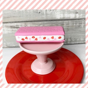 May include: A pink and white ice cream sandwich with red heart sprinkles on a pink cake stand and a red plate.