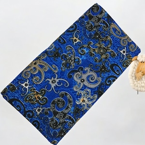 May include: A rectangular blue fabric with a repeating pattern of black, gray, and gold swirling floral designs. The fabric is folded and appears to be a scarf or a piece of cloth. The background is white.