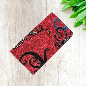 May include: A rectangular red fabric with a floral pattern and black, swirling designs. The fabric has a detailed, ornate design with a touch of blue. The fabric is folded and appears to be a textile.