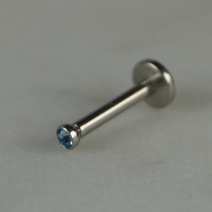 Könnte beinhalten: Ein silberfarbener Lippenpiercing-Stecker mit einem kleinen, runden, hellblauen Edelstein am Ende des Stifts. Der Stecker hat eine flache, kreisförmige Rückseite und einen geraden Stift.