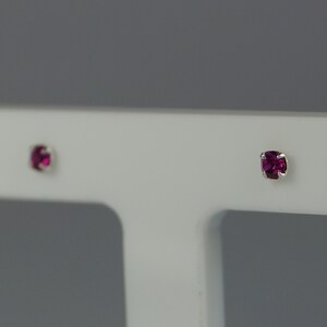 Puede incluir: Un par de pequeños pendientes de botón con piedras preciosas de color magenta y corte cuadrado. Las piedras están engastadas en metal plateado. Los pendientes se muestran sobre un fondo blanco.