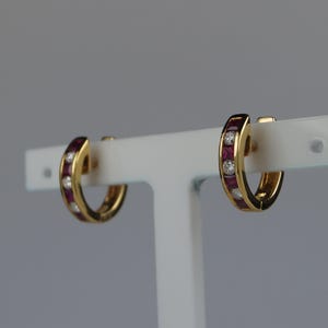 May include: A pair of gold hoop earrings featuring a channel setting with alternating square-cut red rubies and round-cut white diamonds. The earrings are displayed on a white T-shaped stand, showcasing the polished metal finish and the vibrant gemstone arrangement.
