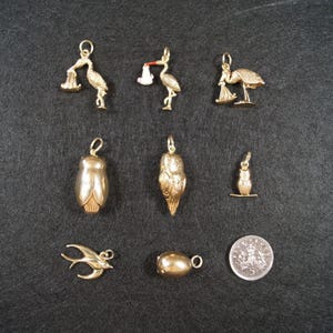 May include: A collection of nine gold-toned metal charms including three stork figures, two owl figures, a small bird, a swallow, and a smooth oval bead, accompanied by a circular silver-colored coin with a crown emblem and the number 5.
