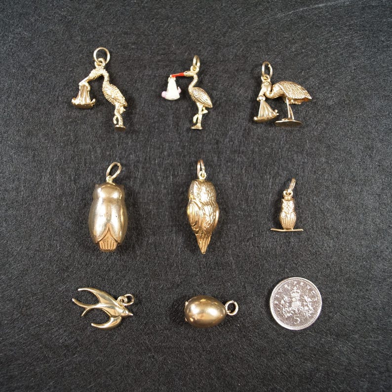 May include: A collection of nine gold-toned metal charms including three stork figures, two owl figures, a small bird, a swallow, and a smooth oval bead, accompanied by a circular silver-colored coin with a crown emblem and the number 5.