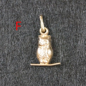 May include: A gold-toned metal owl charm pendant featuring a small owl perched on a horizontal bar with a jump ring at the top for attachment. The letter F is printed in red to the left of the owl charm, which has detailed feather textures.