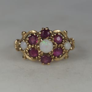 May include: A gold ring with a cluster of gemstones. The ring features a central white opal, surrounded by red rubies and smaller white opals. The band has an ornate, textured design. The ring's design is floral.