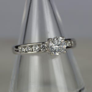 May include: A silver ring with a large, round, clear gemstone as the centerpiece. Smaller, clear gemstones are set along the band. The ring is displayed on a clear, conical stand, showcasing its design and sparkle.