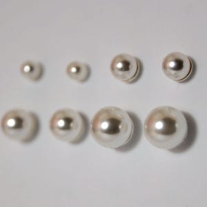 May include: A selection of faux pearl stud earrings in different sizes. The earrings are a creamy white colour with a glossy finish, arranged in two rows against a white background. The earrings are round and have a classic design.