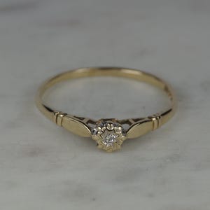 May include: A gold ring featuring a central, small, round, clear gemstone set within a star-shaped, white-toned metal setting. The band is smooth and yellow gold, with decorative, geometric, cut-out shoulders leading to the central stone.