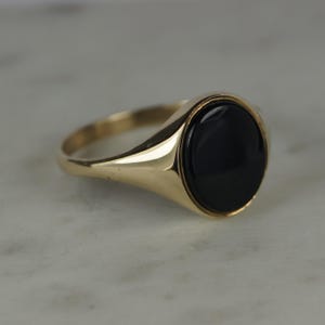 May include: A gold signet ring with a large, oval black stone. The ring has a smooth, polished band and a classic design. The black stone is set in a bezel setting, creating a sleek and elegant look. This ring is a timeless piece of jewelry.