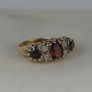 May include: A gold ring featuring a central oval-cut deep red garnet flanked by two smaller round-cut garnets on the outer edges. Four small, round, iridescent white opals are set between the garnets, creating a multi-stone arrangement on a band.
