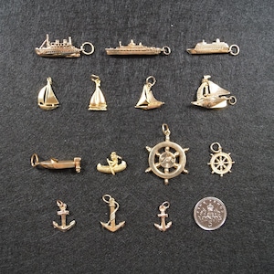 May include: A collection of gold-toned metal charms featuring nautical themes, including various ships, sailboats, a canoe, a ship's wheel, and anchors. A circular coin with the number 5 is included for scale, measuring approximately 0.9 inches in diameter.