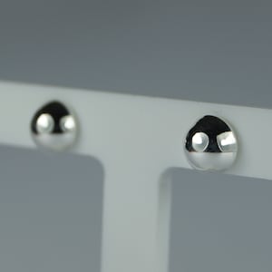May include: Two small, shiny, silver stud earrings. Each earring has a rounded, teardrop shape with a reflective surface. The earrings are displayed against a white background.