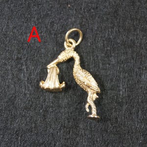 May include: A gold-colored metal charm in the shape of a stork carrying a bundle in its beak. The stork is depicted standing on one leg with textured feather details across its body. A small circular jump ring is attached to the top of the stork.